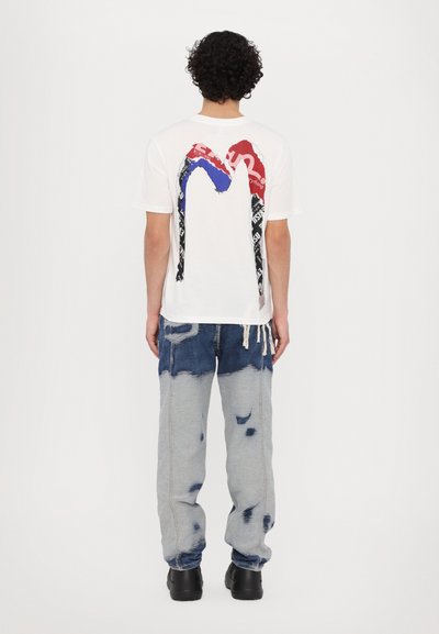 T-shirt blanche avec un graphique rouge et bleu au dos, associée à un pantalon en denim clair déchiré et des chaussures noires.
