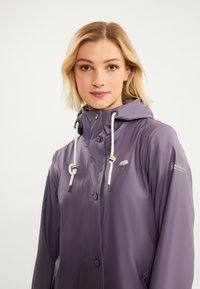 Violeta lietus jaka ar kapuci, baltām auklām un spiedpogām. Dizains ietver augstākā līmeņa samazināšanas lietussarga logotipu un sānu kabatas.