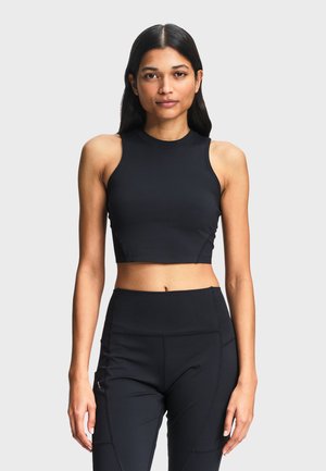 Femme aux longs cheveux foncés portant un crop top noir sans manches et un legging noir taille haute, debout devant un fond gris clair uni.