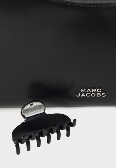 Sac en cuir noir avec une texture lisse, arborant un logo en argent. Un clip à cheveux noir avec 10 dents est placé à l'avant.