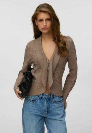 Femme aux longs cheveux ondulés portant un cardigan marron en tricot et un jean bleu, tenant une pochette noire avec une main dans sa poche.