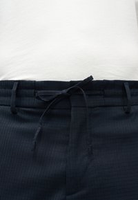 Gros plan sur une taille portant un pantalon bleu marine à motif quadrillé subtil avec un cordon de serrage en tissu noué sur une chemise blanche.