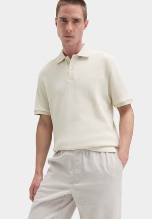 Młody mężczyzna ubrany w kremową, teksturowaną koszulkę polo z krótkim rękawem i jasnobeżowe spodnie, stojący z jedną ręką w kieszeni na jednolitym tle.