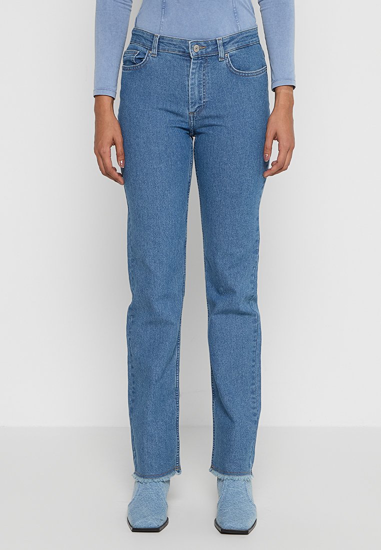 Persoon in rechtgesneden blauwe jeans met rafelige hemden en lichtblauwe puntige laarzen, staande tegen een witte achtergrond.