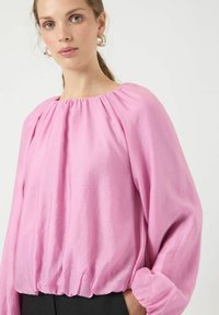 Rosa bluse med lange ærmer, kropsnært snit ved halsen og elastisk kant forneden. Fremstillet af let vævet stof med løse, flagrende ærmer og en glat tekstur.