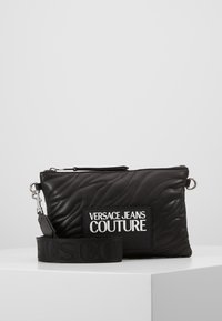 Svart kviltad läderclutch med en stor logopatch, metalliska accenter och ett avtagbart band med präglad text.