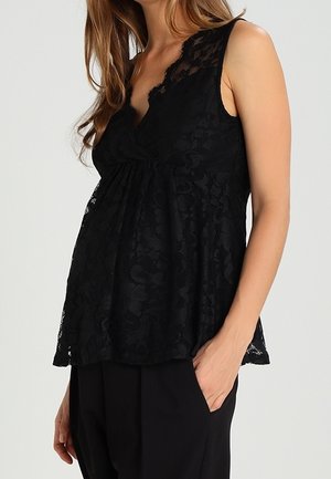 Gravid kvinde iført en ærmeløs sort blondetop til graviditet med V-hals og sorte bukser, stående med den ene hånd i lommen.