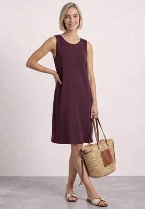 Femme aux cheveux blonds courts portant une robe violette sans manches, tenant un sac tissé, chaussée de sandales beige, debout sur un sol gris devant un fond uni.