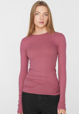 Jeune femme aux cheveux blonds raides, portant un haut côtelé rose ajusté à manches longues et un pantalon noir, debout devant un fond uni.