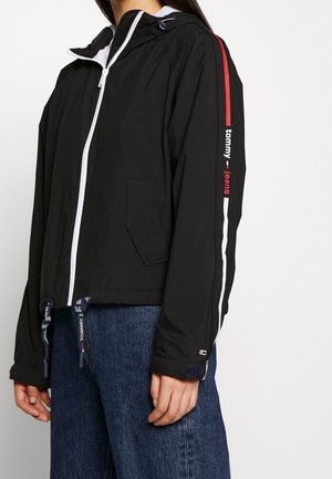 Persoon die een zwarte jas met ritssluiting draagt, met rode en witte strepen en de tekst "tommy jeans" op de mouw, gecombineerd met blauwe spijkerbroeken.