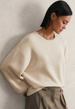 Kremowy sweter dziany o luźnym kroju, z prążkowanym dołem i srebrnym łańcuchem przy dekolcie, w połączeniu z beżowymi spodniami o wysokim stanie i plisami.