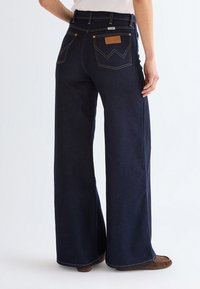 Woman wearing dark blue wide-leg Wrangler jeans with brown moccasin shoes, shown from the back at mid-calf length.