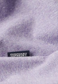 Tessuto lavanda con una consistenza morbida, caratterizzato da un'etichetta nera con testo bianco che recita "SUPERDRY" cucita. Variazione di colore sottile.