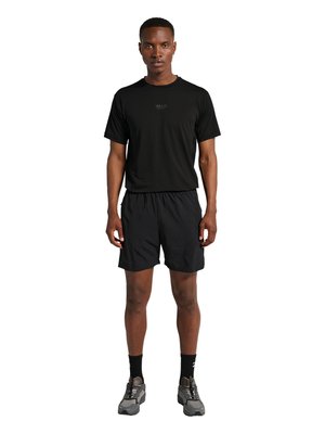 Mann steht frontal, trägt ein schwarzes kurzärmeliges Shirt mit „HALO“-Logo, schwarze Shorts, schwarze Socken und graue Sportschuhe.