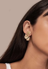 Mujer con cabello largo y oscuro que lleva pendientes de botón en forma de pétalo abstracto texturizado en dorado, sobre un fondo beige.