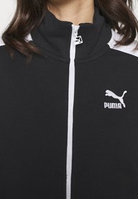 Sort zip-jakke med høj krave, hvide detaljer på skuldrene og et fremtrædende hvidt Puma-logo på venstre bryst.
