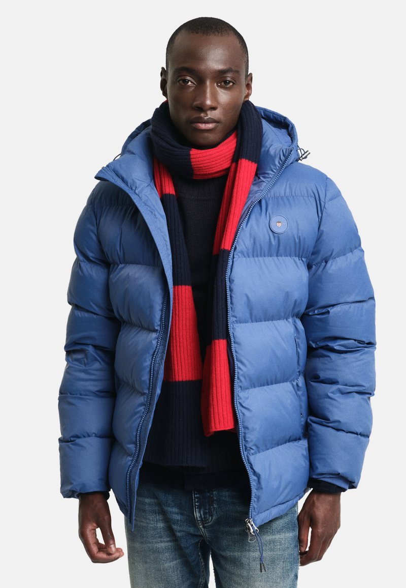 Modrá péřová bunda s kapucí, prošívaným designem a předním zipem. V doprovodu červeno-námořnického pruhovaného šálu. Spodní část s casual džíny.