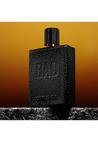 Sort tekstureret rektangulær parfumeflaske med præget "BAD" og "DIESEL", stående på en ru stens overflade mod en gradient mørk orange baggrund.