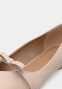 Ballerine rose clair en similicuir lisse, avec une fine bride et un nœud décoratif sur le bout. Intérieur beige.