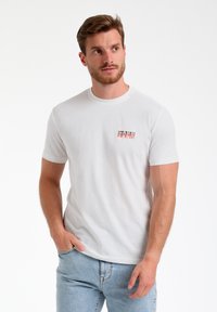 Balta īsām piedurknēm T-kreklā, izgatavotā no kokvilnas, ar apaļu dekoltē. Uz kreisās krūts daļas ir neliels, daudzkrāsains logotips. Brīva piegriezuma.