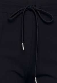 Zwarte sweatpants hebben een tailleband met trekkoord en zwarte gevlochten koorden met metalen aglets, en tonen een soepele, zachte textuur.