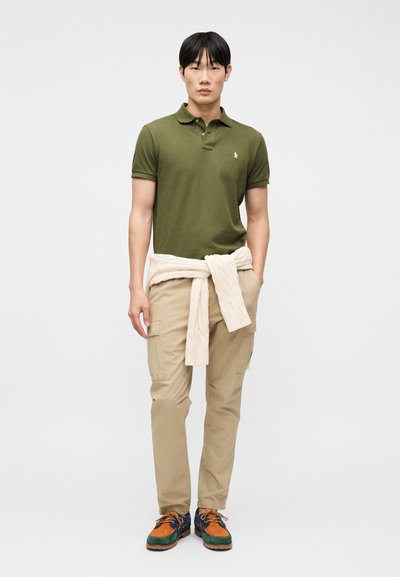 Άνδρας που φοράει ελιά πράσινη πόλο μπλούζα, μπεζ παντελόνι καρό, κρεμ πουλόβερ δεμένο γύρω από τη μέση και πολύχρωμα παπούτσια με κορδόνια, στέκεται όρθιος.