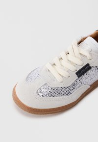 Zapatillas deportivas de ante blancas con cordones beige, paneles laterales de malla, detalles negros y suela de goma marrón sobre un fondo claro.