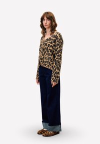 Vrouw die staat, gekleed in een luipaardprint cardigan en bijpassende slippers, met een wijde, omgeslagen donkerblauwe spijkerbroek, tegen een effen lichte achtergrond.