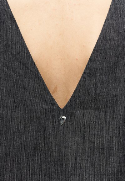 Κοντινή λήψη ενός σκούρου ντένιμ ρούχου με βαθιά λαιμόκοψη σε σχήμα V που δείχνει γυμνό δέρμα και μια μικρή ασημένια παραμάνα που συγκρατεί το ύφασμα στο κάτω μέρος.