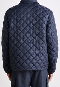 Veste matelassée bleu marine présentant un motif en losanges, une texture en tissu et un col classique, avec une coupe droite et un design minimaliste.