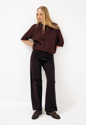 Žena s dlhými blond vlasmi stojí opretá o bielu stenu, oblečená v tmavohnedom prehnanom svetri, širokých čiernych nohaviciach a tmavohnedých topánkach.