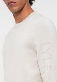 Világos bézs crew neck pulóver kötött anyagból. Bordás gallérral és textúrázott "LYLE" részlettel a ujjakon rendelkezik. Simább felület.