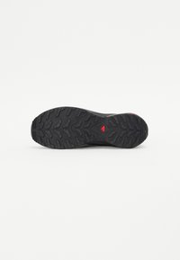 Semelle de chaussure en caoutchouc noir avec un motif de semelle texturé pour l'adhérence, avec un accent triangulaire rouge proéminent et des bords lisses.