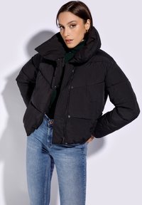 Chaqueta puffer negra con cuello alto, diseño acolchado y cremallera frontal. Combinada con vaqueros azules, que tienen una silueta ajustada.