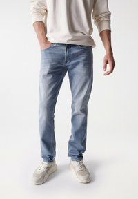 Jeans in denim azzurro con effetto sbiadito, caratterizzati da un design a cinque tasche, abbinati a sneakers color crema e a un maglione beige.