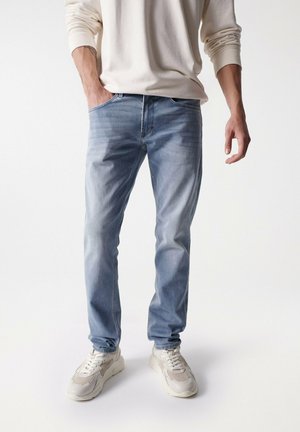 Jeans de denim azul claro con un lavado desvanecido, con un diseño de cinco bolsillos, combinados con zapatillas color crema y un suéter beige.