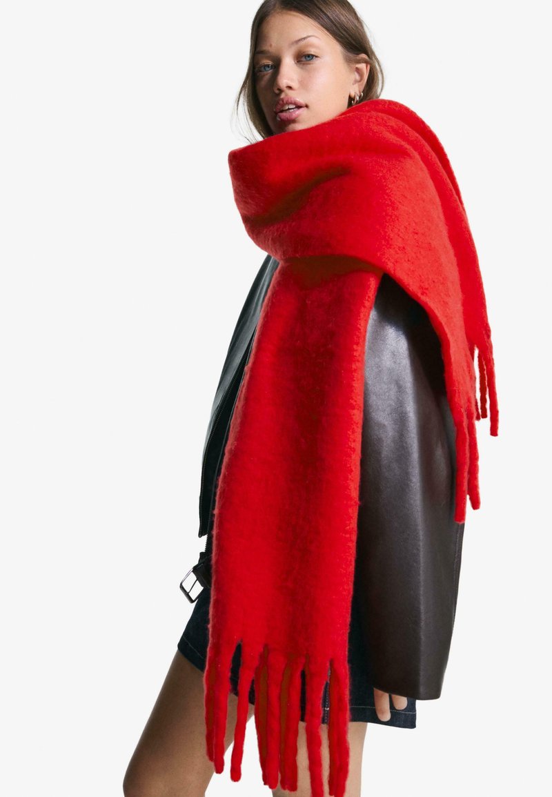 Fiatal nő, aki egy nagy, élénkpiros rojtokkal díszített sálat visel egy fekete bőrkabát felett és sötét farmer szoknyában, a vállán át a kamerába néz.