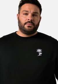 Sudadera negra con cuello redondo y un diseño de palmera bordado en blanco en el lado izquierdo del pecho. Textura suave, ajuste casual.