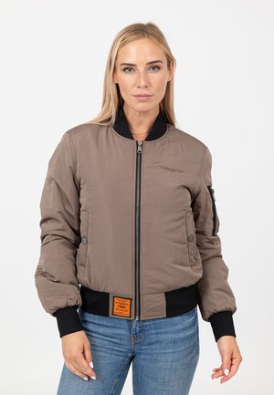 Woman wearing a taupe bomber jacket with black collar and cuffs, front zipper, patch on hem, and blue jeans, standing against a white background.