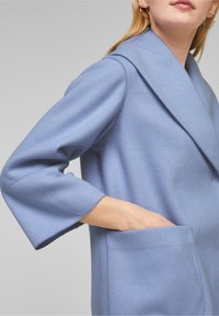 Ljusblå kappa i mjukt texturerat tyg, med bred krage, trekvartslånga ärmar och en framficka på ena sidan.