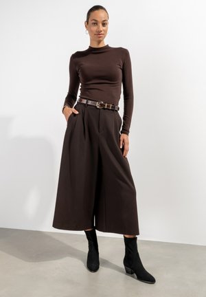 Bruine top met lange mouwen gecombineerd met hooggewaste, wijde bruine broek, aangevuld met een bruine riem en enkellange zwarte laarzen.