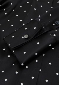 Must valge täpilise mustriga särk, millel on nööbitav mansett ja sile, satiinilaadne tekstuur. Aktsendina üks ümar nööp ja nööpaugud.