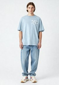 T-shirt oversize azzurro chiaro con testo grafico bianco "Mi Amor" e disegno a forma di cuore, abbinato a jeans in denim blu sbiadito e sneakers bianche.