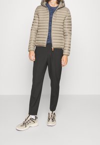 Beige doorgestikte gewatteerde jas met capuchon, marineblauw gestructureerd overhemd, zwarte taps toelopende broek en beige sportieve schoenen met zwarte accenten.