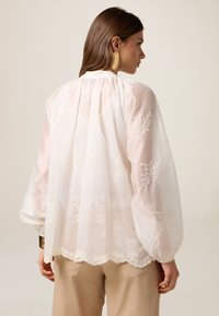 Blusa blanca bordada con mangas abullonadas, dobladillo festoneado y un cuello alto. Presenta delicados patrones florales en tela translúcida.