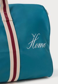 Borsa teal con manico a righe crema e rosso, con la parola "Home" ricamata in corsivo sul lato.