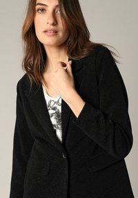 Zwarte blazer met een gestructureerde afwerking, voorzien van een enkele knoopsluiting en twee voorzakken. Draagt over een witte graphic tee.