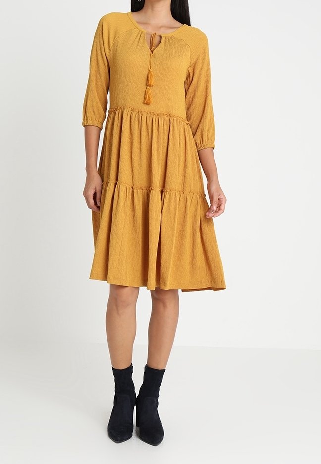 Femme portant une robe moutarde texturée à volants, longueur genou, manches 3/4, col à nouer avec pompons, et bottines noires.