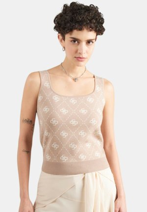 Donna con capelli corti e ricci, indossa un top beige senza maniche con motivo e una gonna color crema, in piedi contro uno sfondo chiaro e semplice.