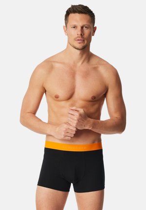 Schwarze Boxershorts mit einem orangefarbenen elastischen Bund, der den Markennamen in heller Schrift zeigt, Modell ohne Hemd dargestellt.